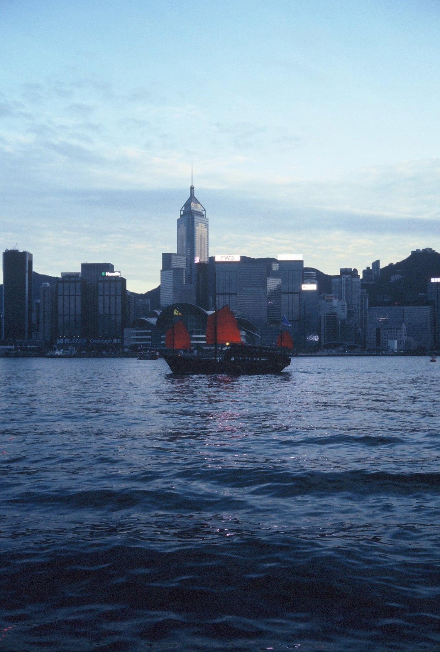 A ferry docked at a harbor at dusk, with the city skyline in the background, captured on KOMICHROME 100 slide film with medium grain.