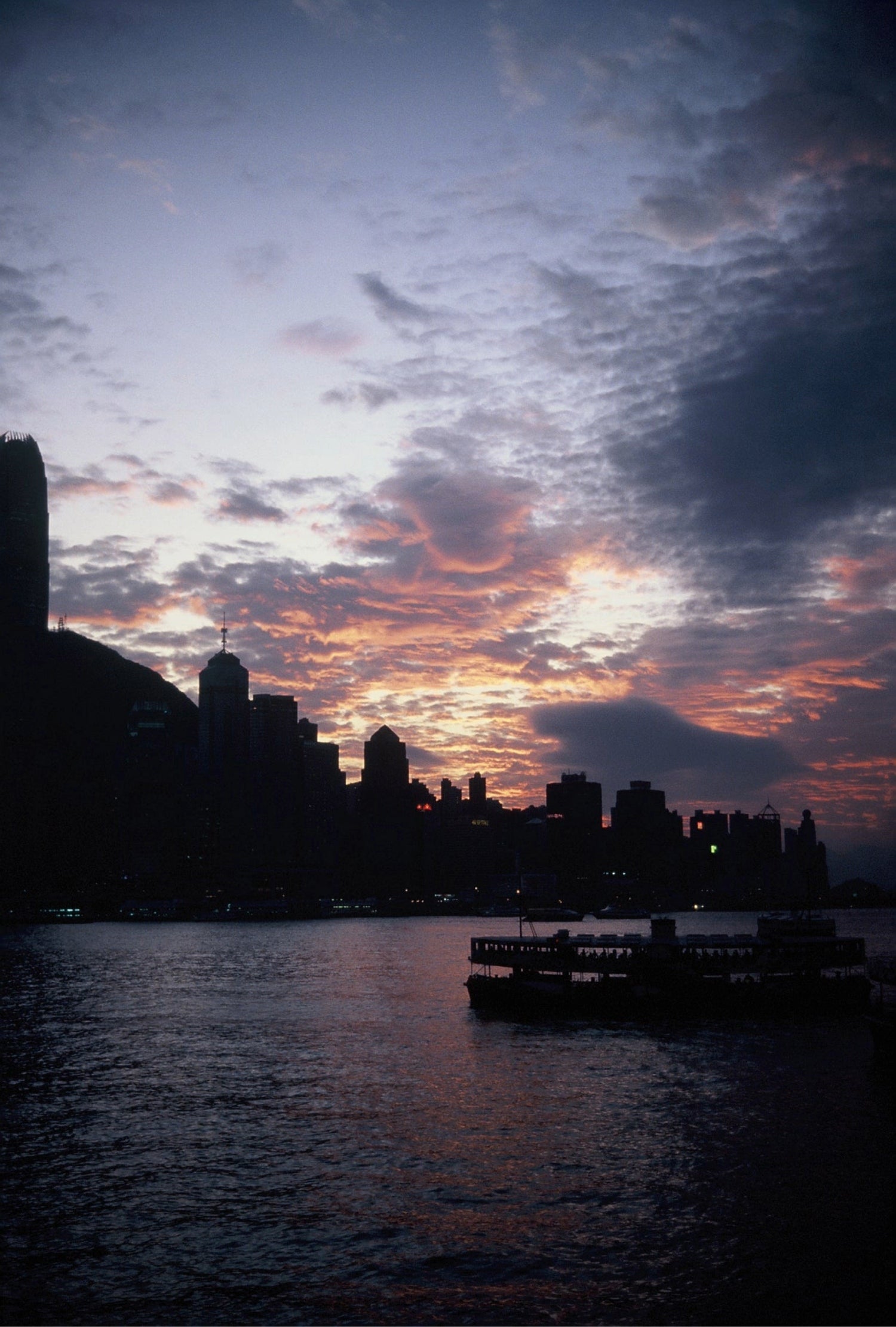 A traditional boat with red sails sailing at sunset against the city skyline, captured on KOMICHROME 100 slide film with medium grain.